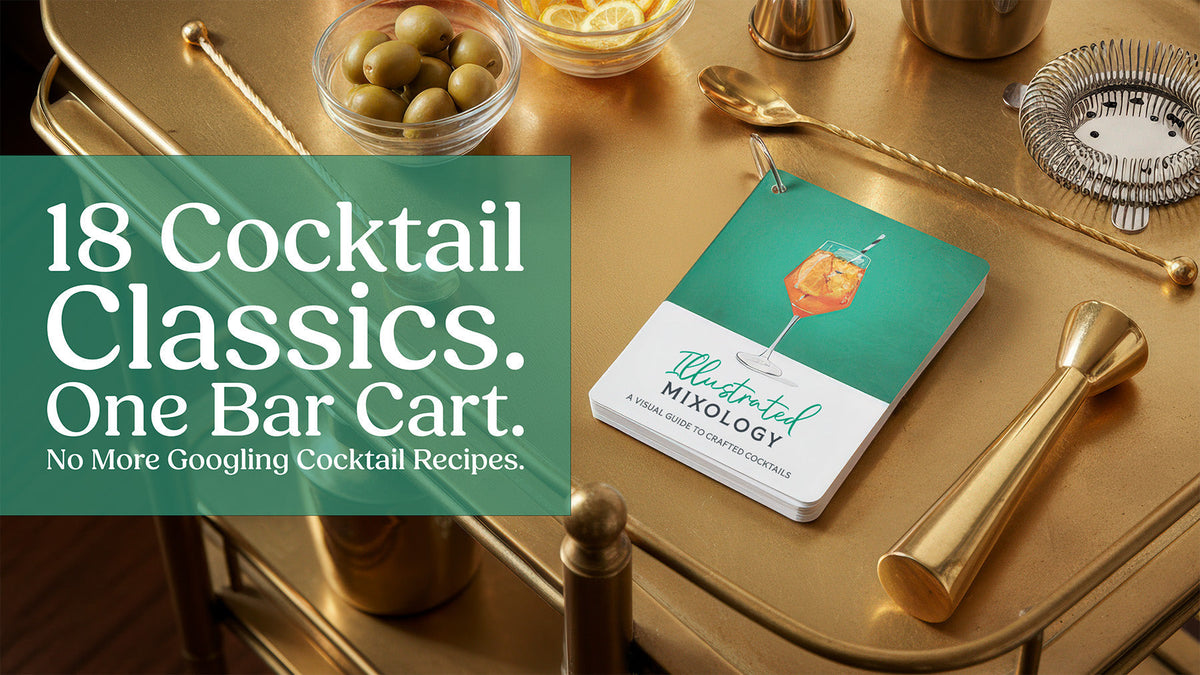 18 cocktail classics, one bar cart. The Illustrated Mixology card deck on a gold bar cart with a jigger, bar spoon, strainer and bowls of olives and lemon slices.