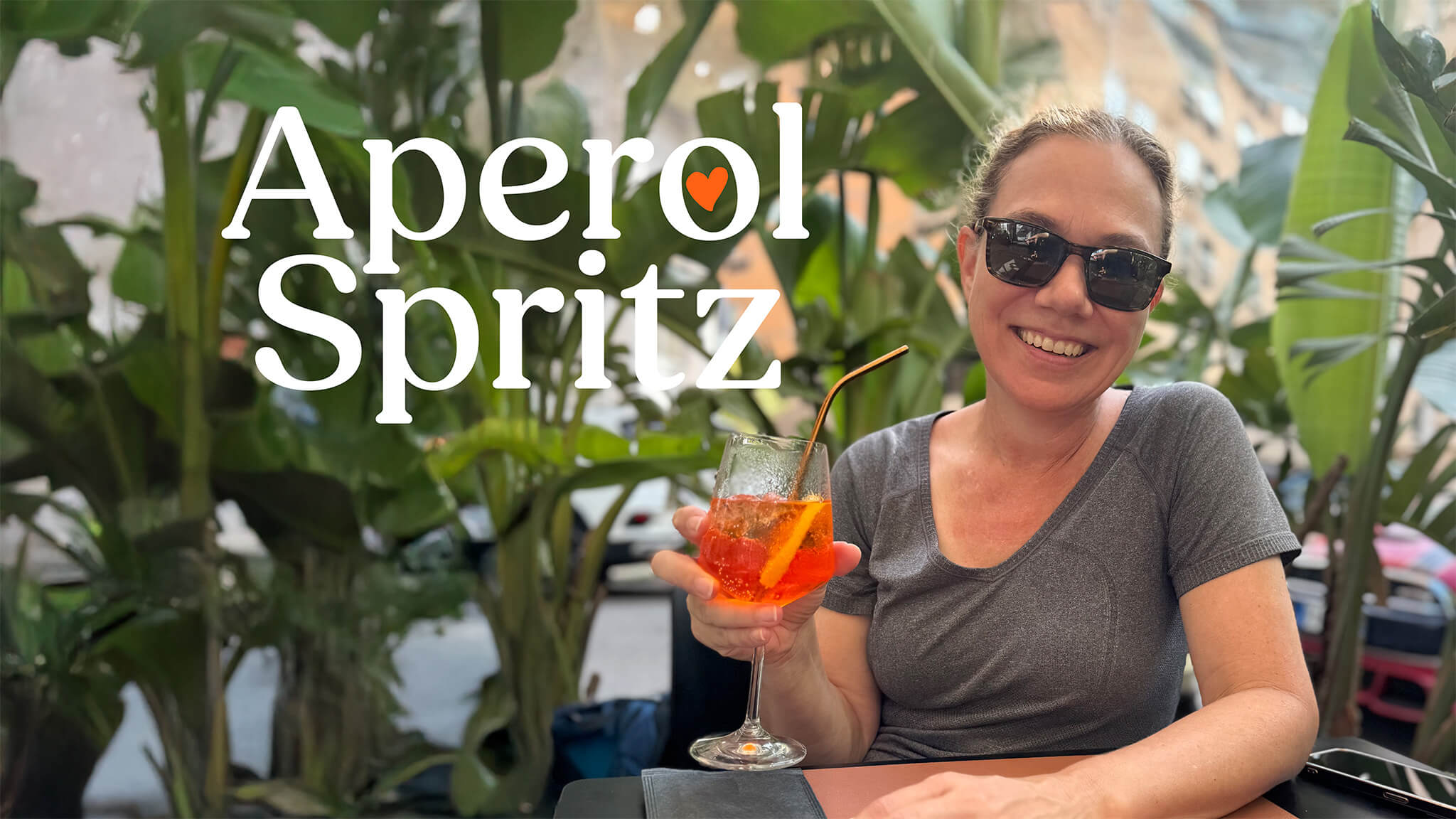 Woman holding an Aperol Spritz cocktail at an outdoor cafe surrounded by tropical plants, with 'Aperol Spritz' text overlay.