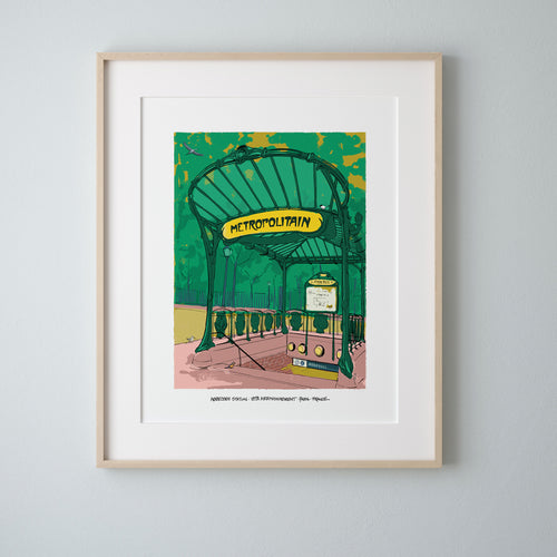 A framed art print of the Abbesses Metro entrance in Paris, matted in white with a light wood frame, hung on a light gray wall.