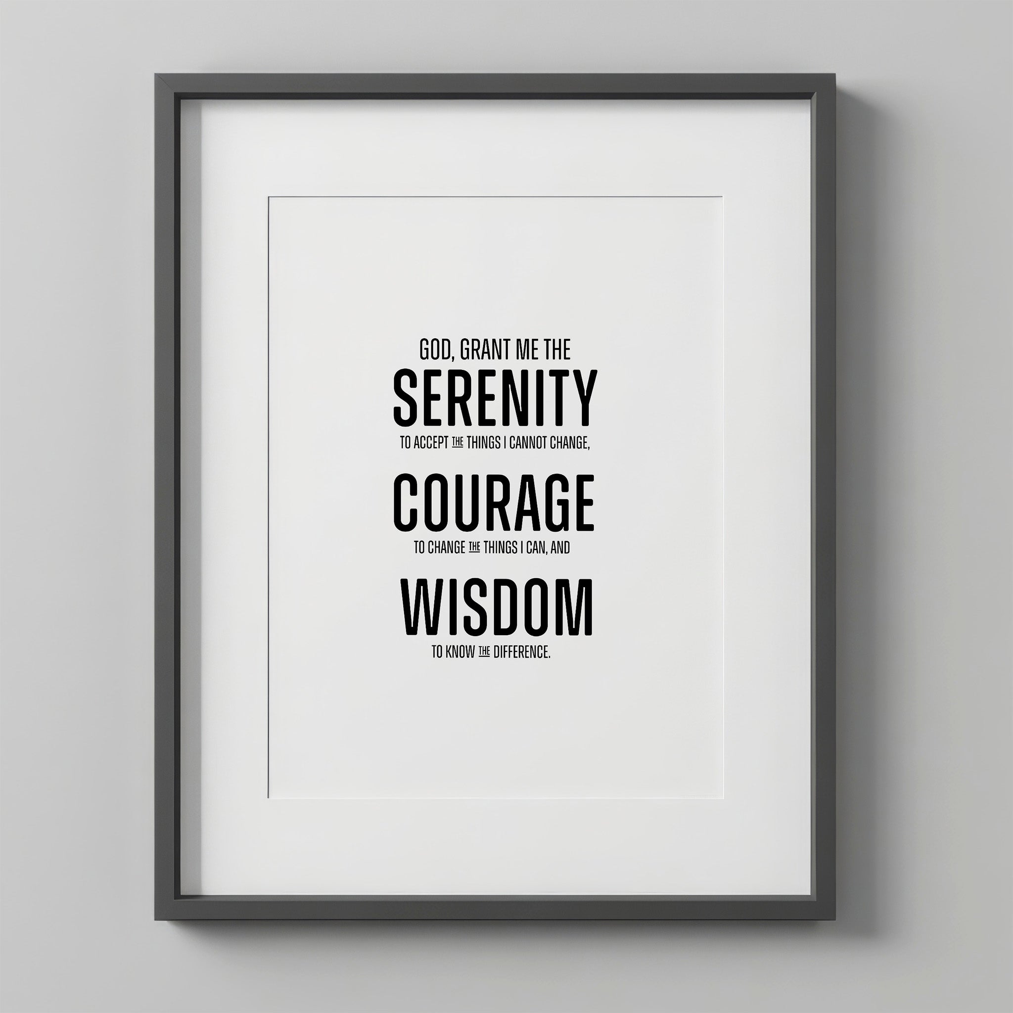 Serenity Prayer Wall Art print in a charcoal-gray frame with a white mat, hung on a light gray wall. The prayer is set in bold modern block type, with SERENITY, COURAGE, and WISDOM in large caps.