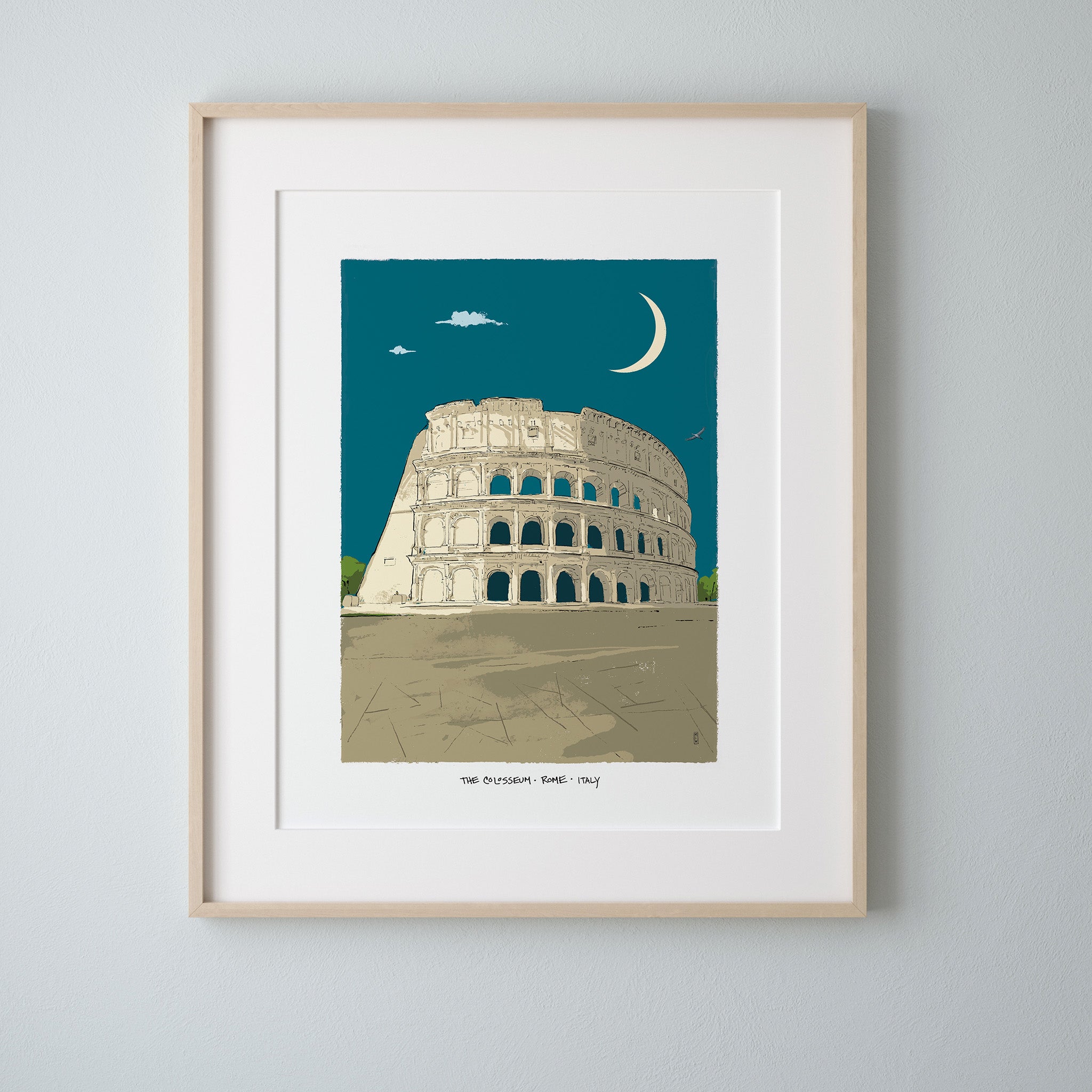 A framed art print of the Colosseum in Rome under a crescent moon, featuring a cream-colored building against a teal sky, matted in white with a light wood frame, hung on a light gray wall.