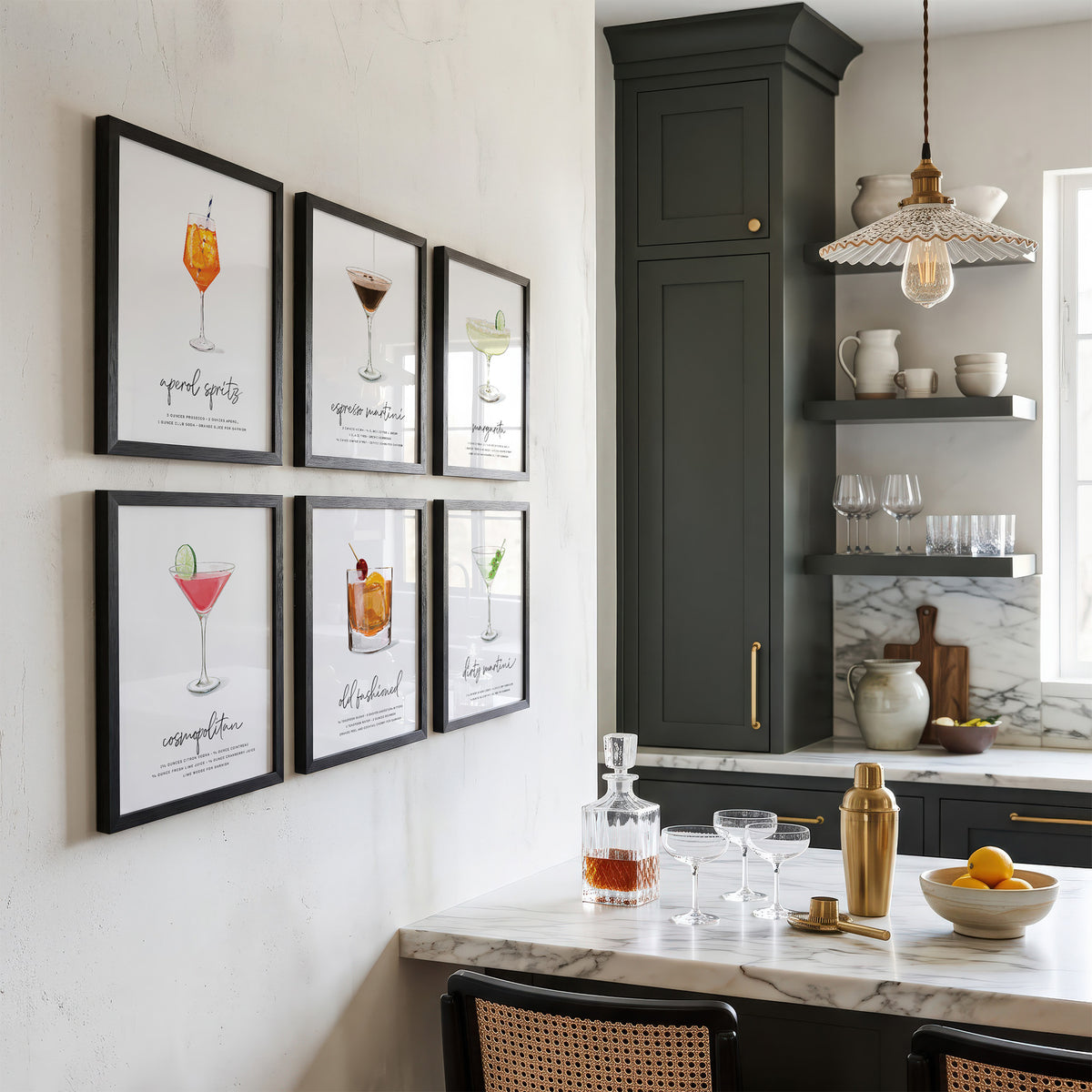 Six framed cocktail art prints with black frames are displayed in a grid on a white wall above a kitchen bar counter. The kitchen features dark green cabinetry, marble countertops, and a hanging glass pendant light.