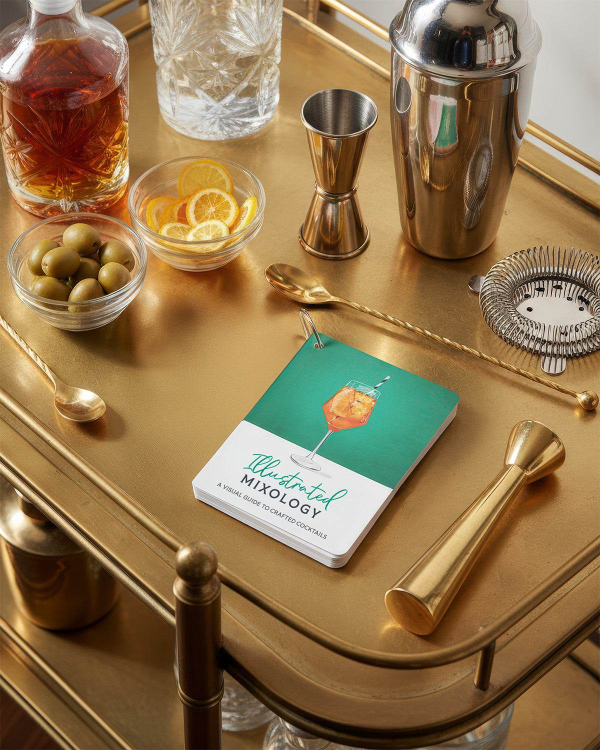 Bartending tools and ingredients on a gold tray with a set of Illustrated Mixology cocktail recipe cards.