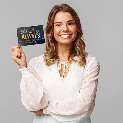 Woman smiling while holding a small 5x7 ‘Love Always, Love Never Fails’ Valentine’s art with a navy background and gold foil accents.