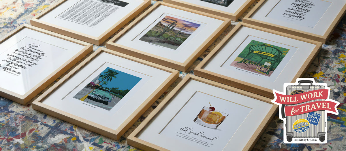 Six framed travel wall rt prints and cocktail wall art with light wood frames and white mats are arranged on a colorful paint-splattered studio drop cloth. 