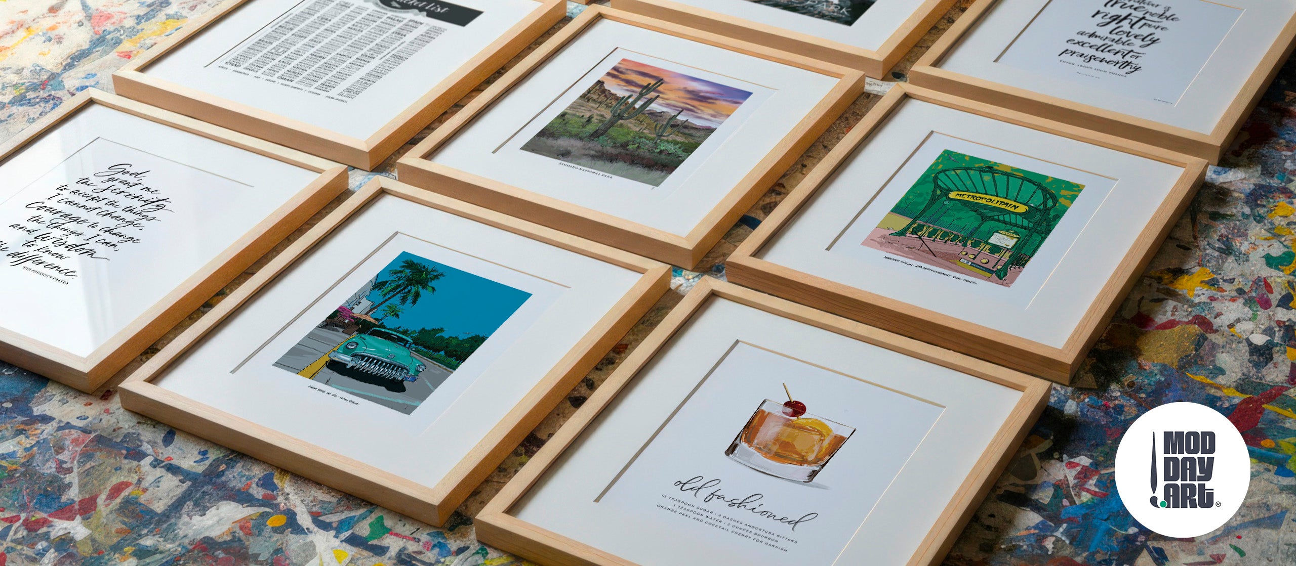 Six Mod Day Art prints in light oak frames with white mats, laid out on a paint-splattered studio floor: the Serenity Prayer, Saguaro National Park, the Abbesses Metro entrance in Paris, Ocean Drive in Miami Beach, and an Old Fashioned cocktail recipe.