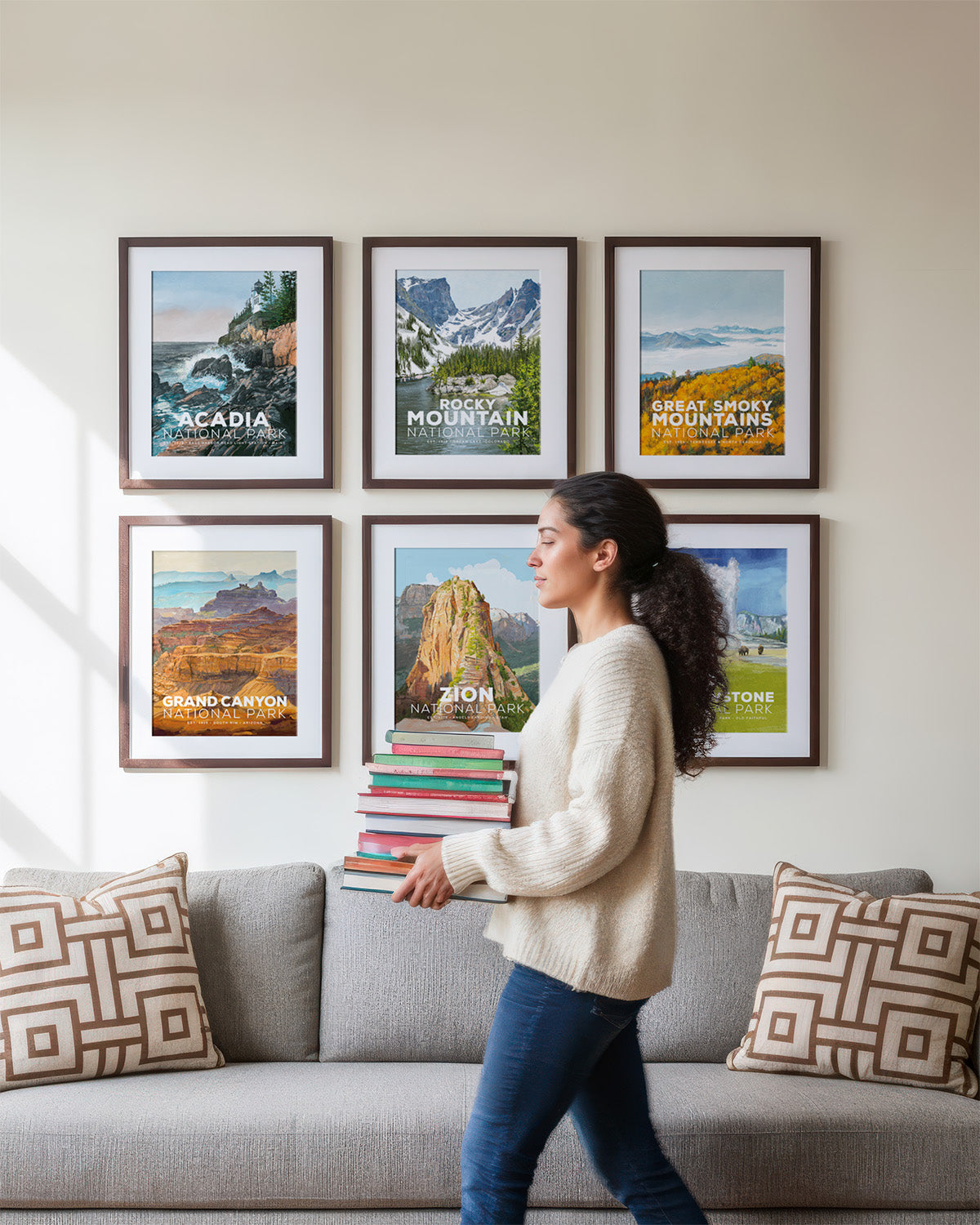Woman carrying stack of books walking past framed Mod Day Art National Parks posters including Acadia, Rocky Mountain, Great Smoky Mountains, Grand Canyon, Zion, and Yellowstone on living room wall.