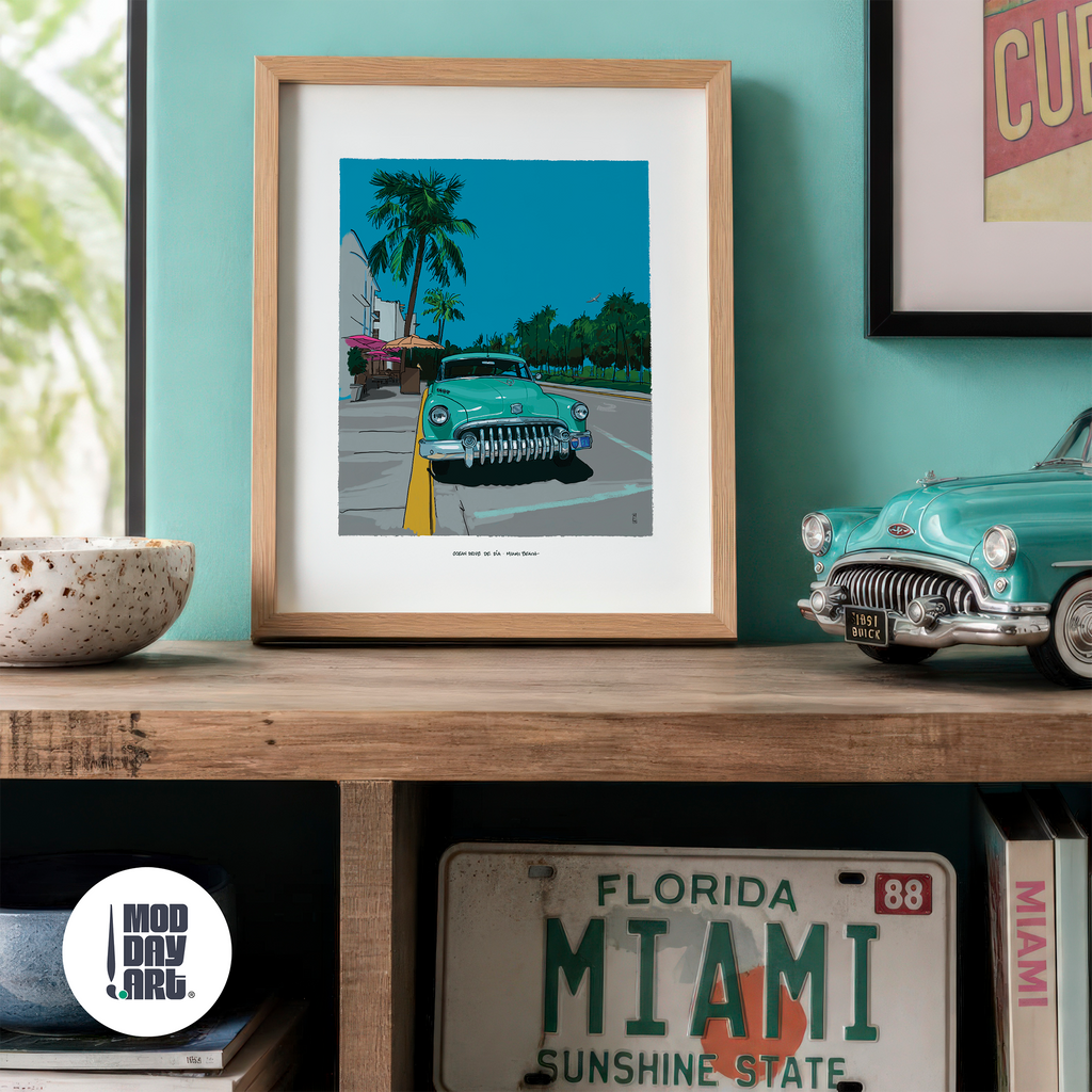 Ocean Drive by Day Miami Beach art print in an oak frame, propped on a wood console against a teal wall beside a model 1951 Buick, a vintage Florida license plate and a stack of books.