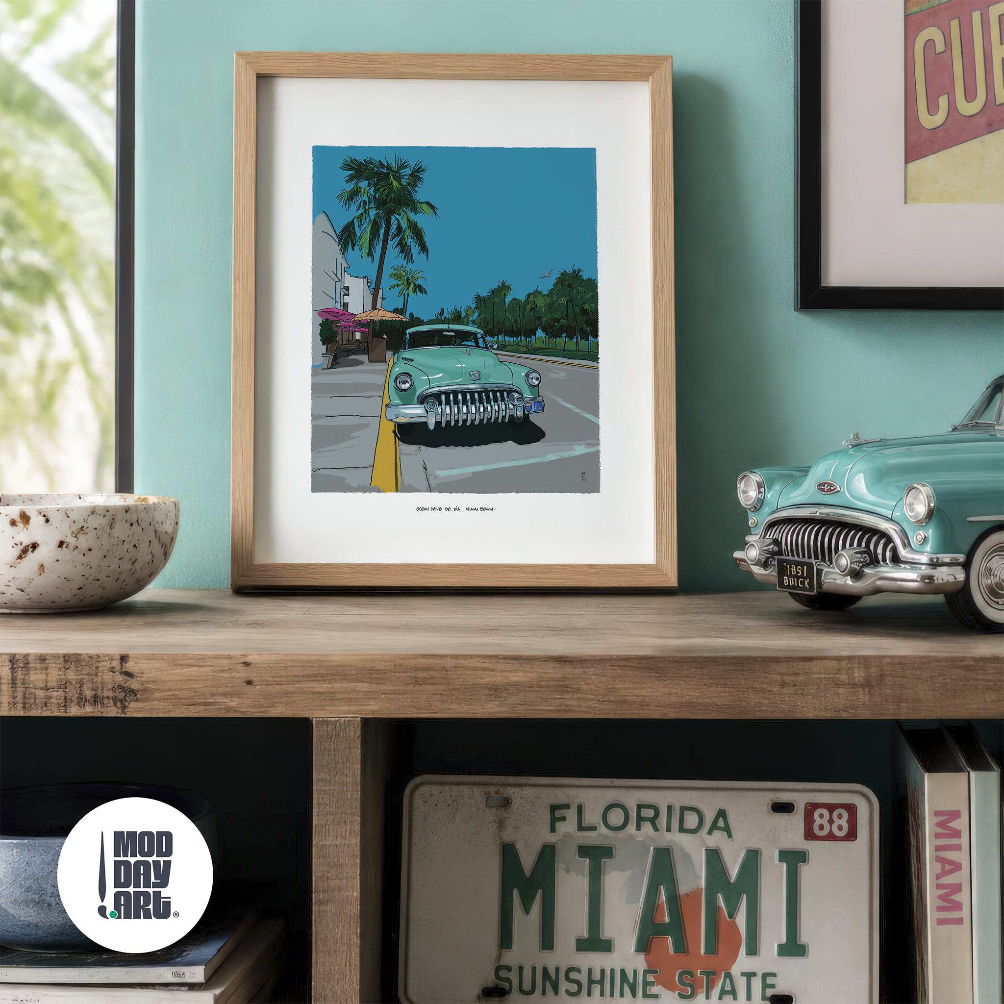 Ocean Drive by Day Miami Beach art print in an oak frame, propped on a wood console against a teal wall beside a model 1951 Buick, a vintage Florida license plate and a stack of books.