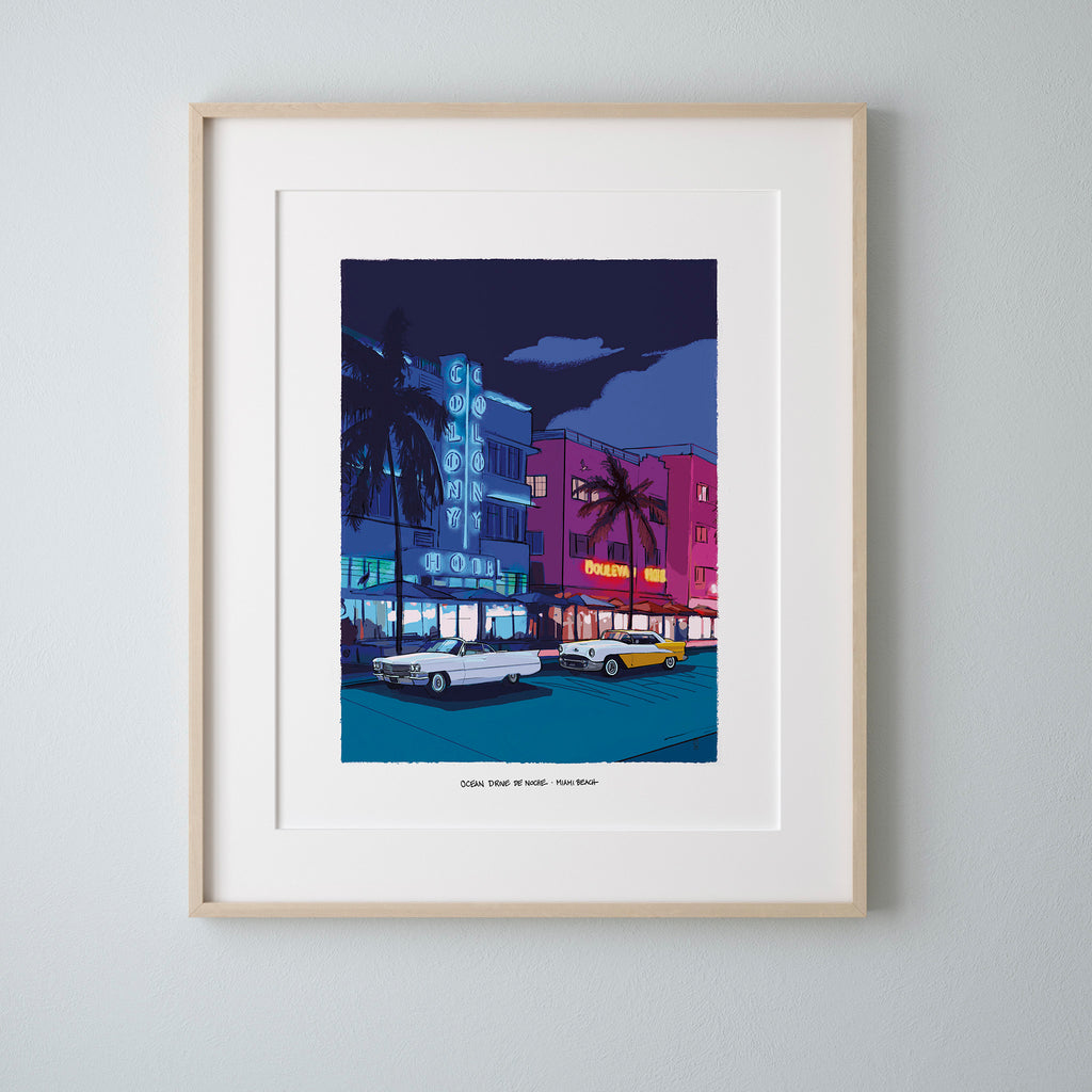A framed art print depicting Ocean Drive in Miami Beach at night, featuring vibrant neon signs, palm trees, and vintage cars. The print is presented in a light natural wood frame with a wide white mat, mounted on a light gray wall.