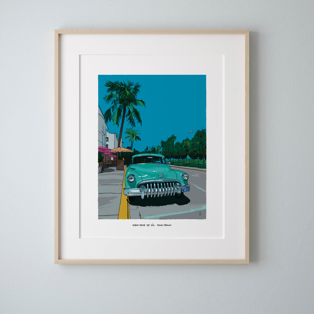 Ocean Drive by Day Miami Beach art print in a light natural wood frame with a wide white mat, hung on a pale gray wall. The hand-illustrated scene shows a mint-green Buick parked at the curb under palm trees on a bright blue morning.