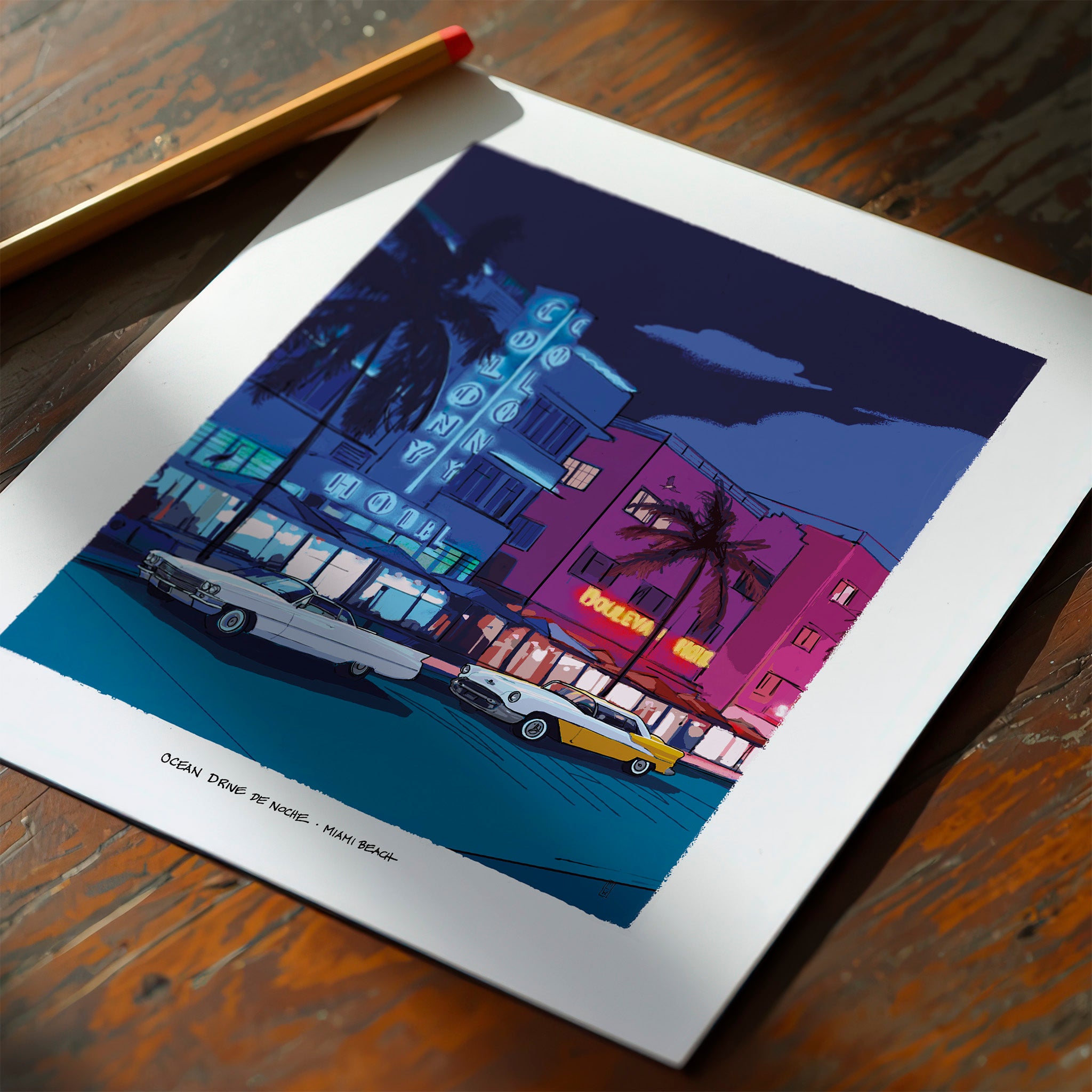 A square art print with a white border lies on a wooden table, showing a stylized nighttime Ocean Drive Miami Beach scene with neon signs, palm trees, and vintage cars. A pencil rests in the upper-left corner of the print.