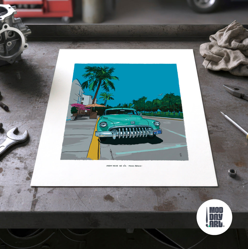 Unframed Ocean Drive by Day Miami Beach art print lying on a garage workbench among hand tools, showing the full sheet with its wide white border and the mint-green Buick illustration.