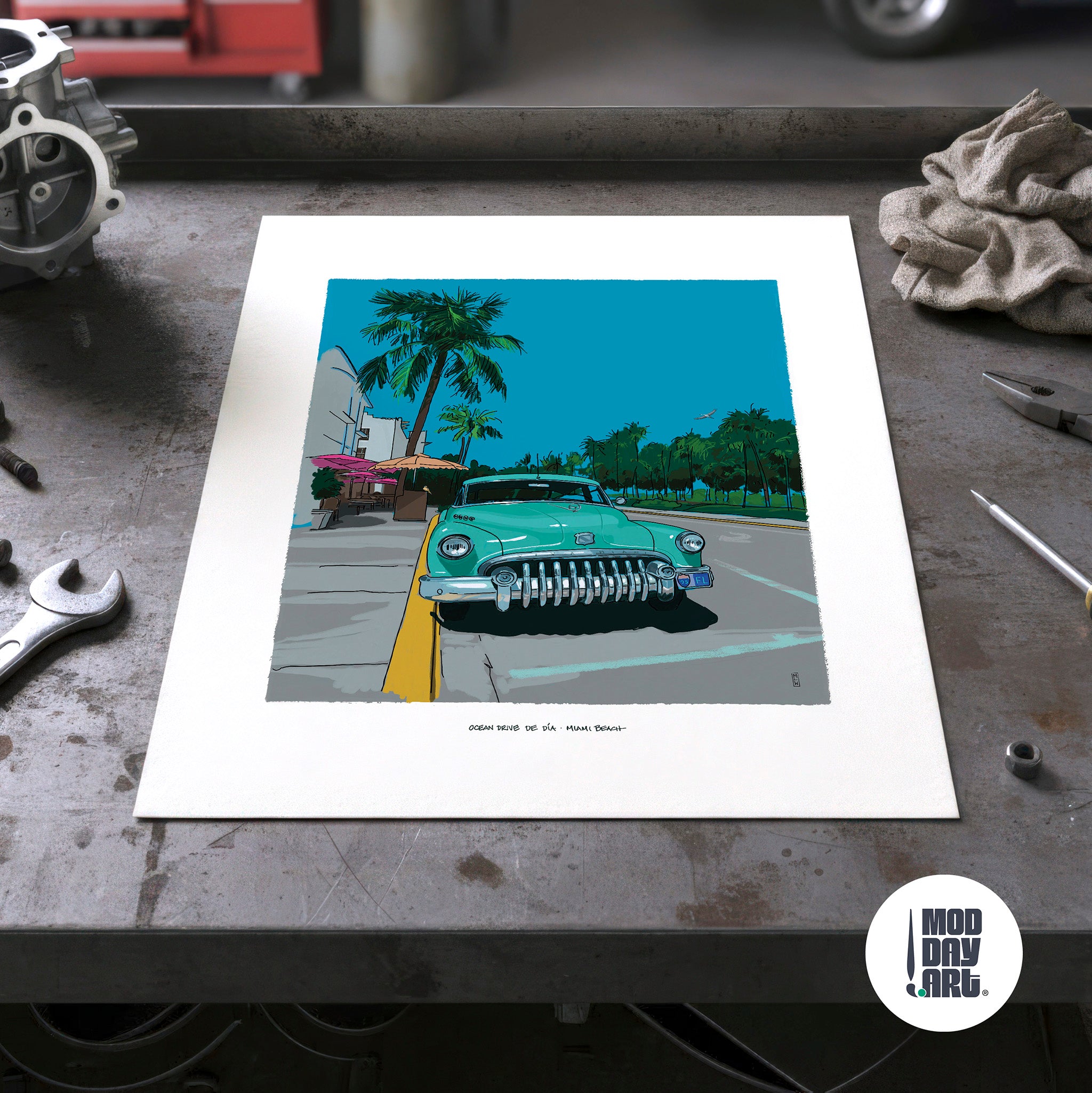 Unframed Ocean Drive by Day Miami Beach art print lying on a garage workbench among hand tools, showing the full sheet with its wide white border and the mint-green Buick illustration.