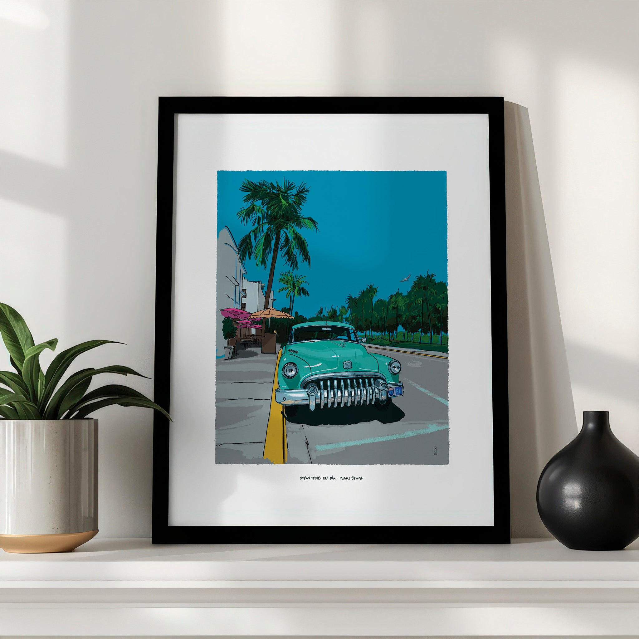 Ocean Drive by Day Miami Beach art print in a black frame with a white mat, leaning on a white shelf between a potted plant and a black vase.