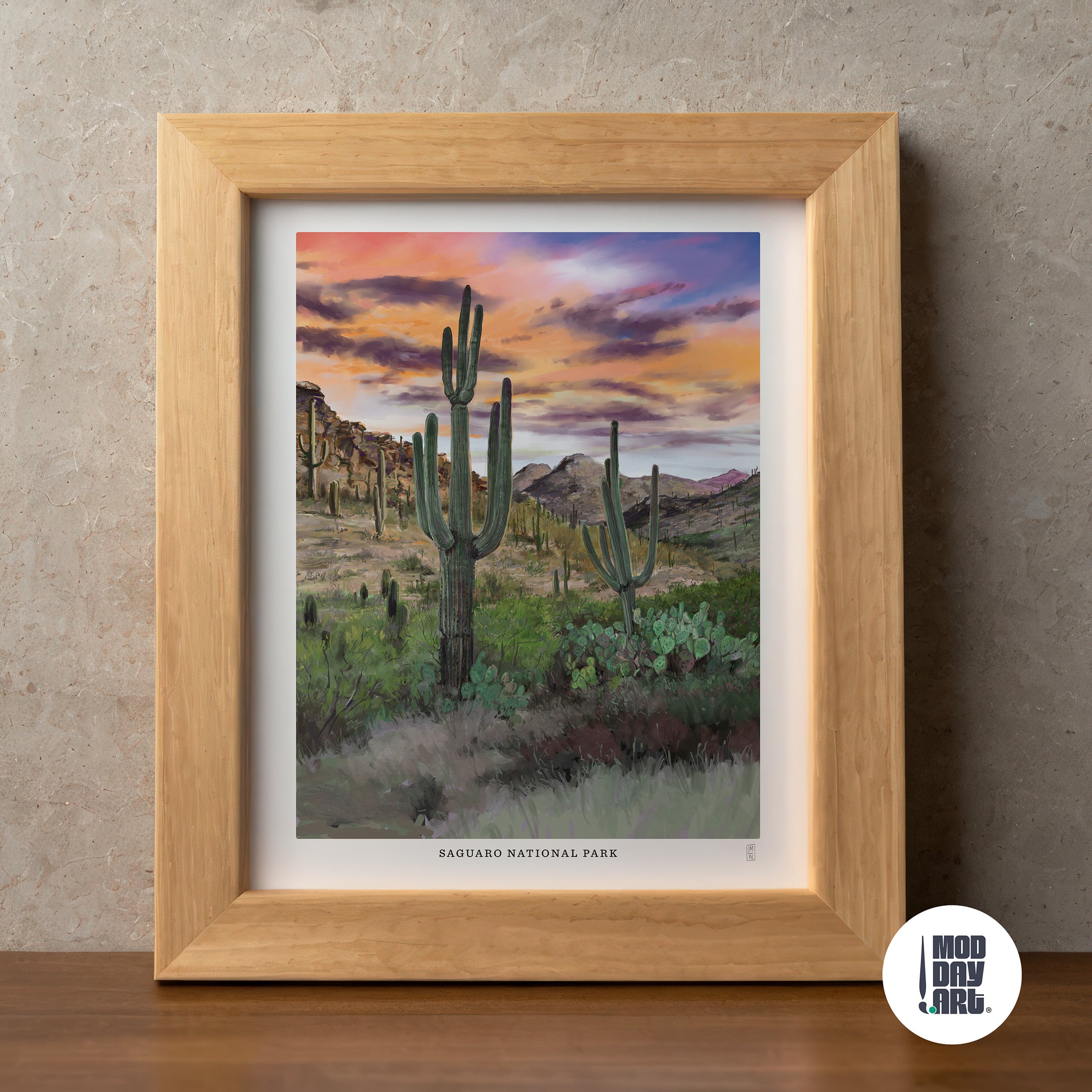 Framed hand-signed Saguaro National Park artwork of a desert landscape with cacti and mountains, displayed on a wooden surface.