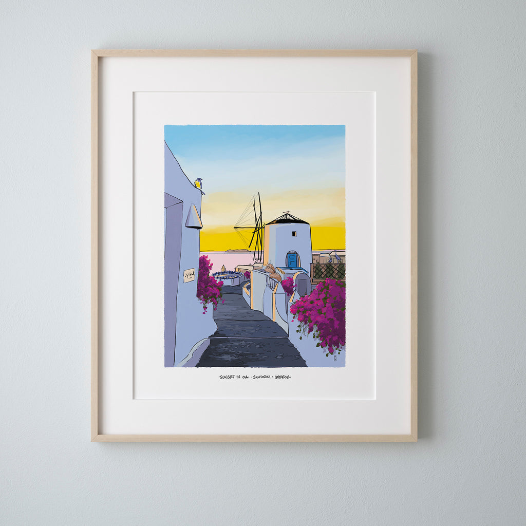 A framed art print depicting a colorful village street in Santorini at sunset, featuring white buildings, purple bougainvillea flowers, and a windmill under a blue and yellow sky. The print is matted in white and housed in a light natural wood frame, mounted on a light gray wall.
