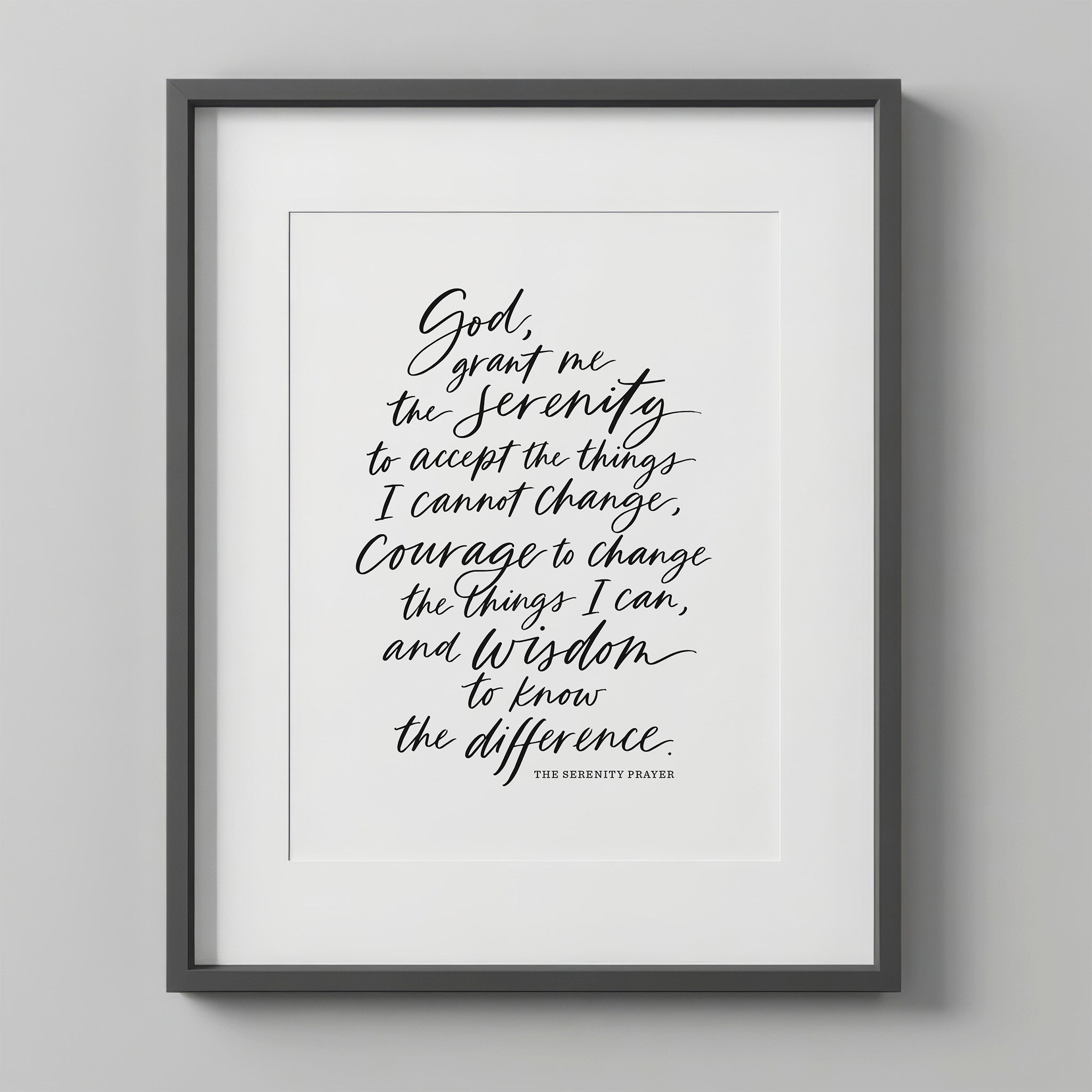 Serenity Prayer Wall Art print in the handwritten script style, black text on white, in a charcoal frame with a white mat on a light gray wall.