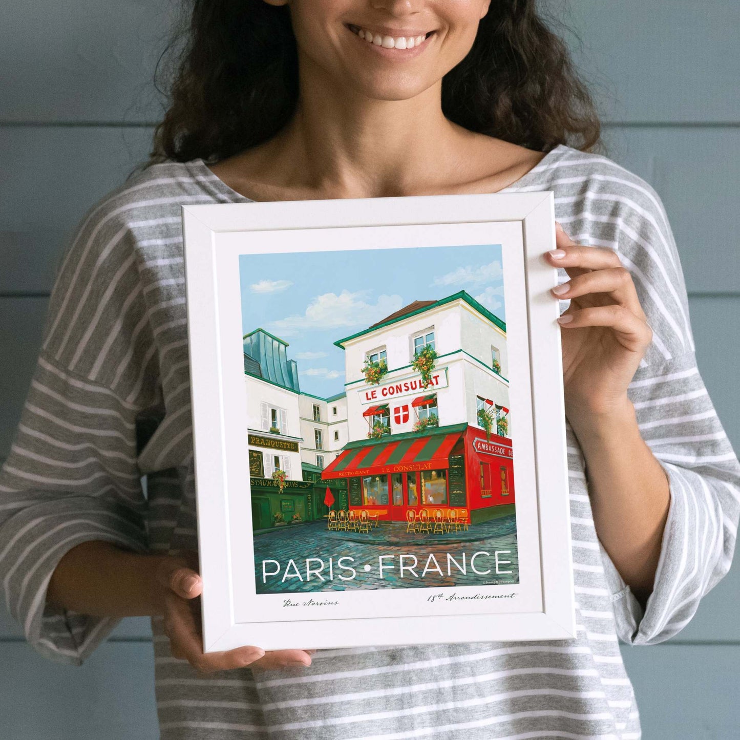 Woman holding Le Consulat Paris Wall Art elegantly displayed in a white frame (frame not included), perfect for adding a Parisian vibe to any space.