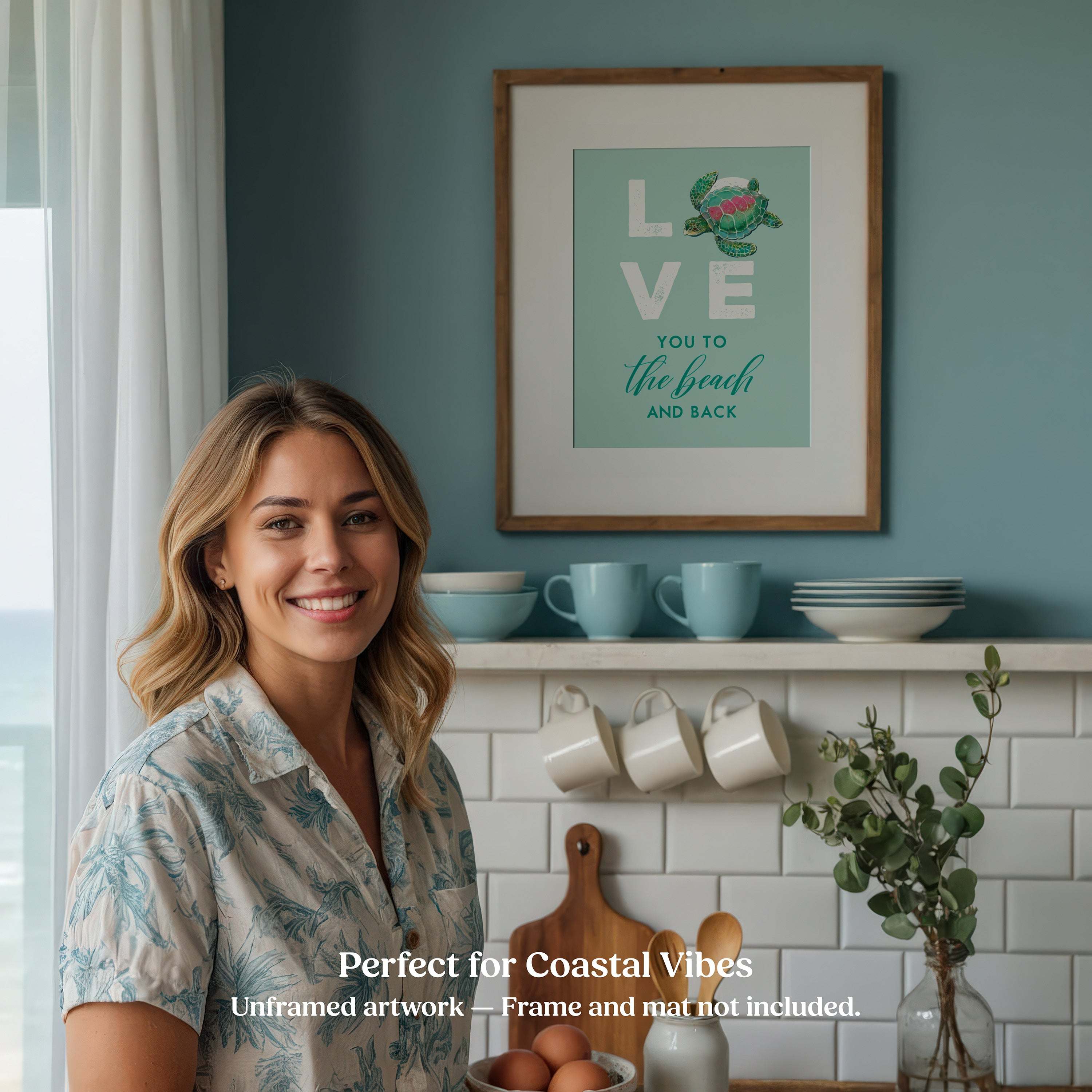 Woman standing in a kitchen with framed coastal beach artwork on the wall.