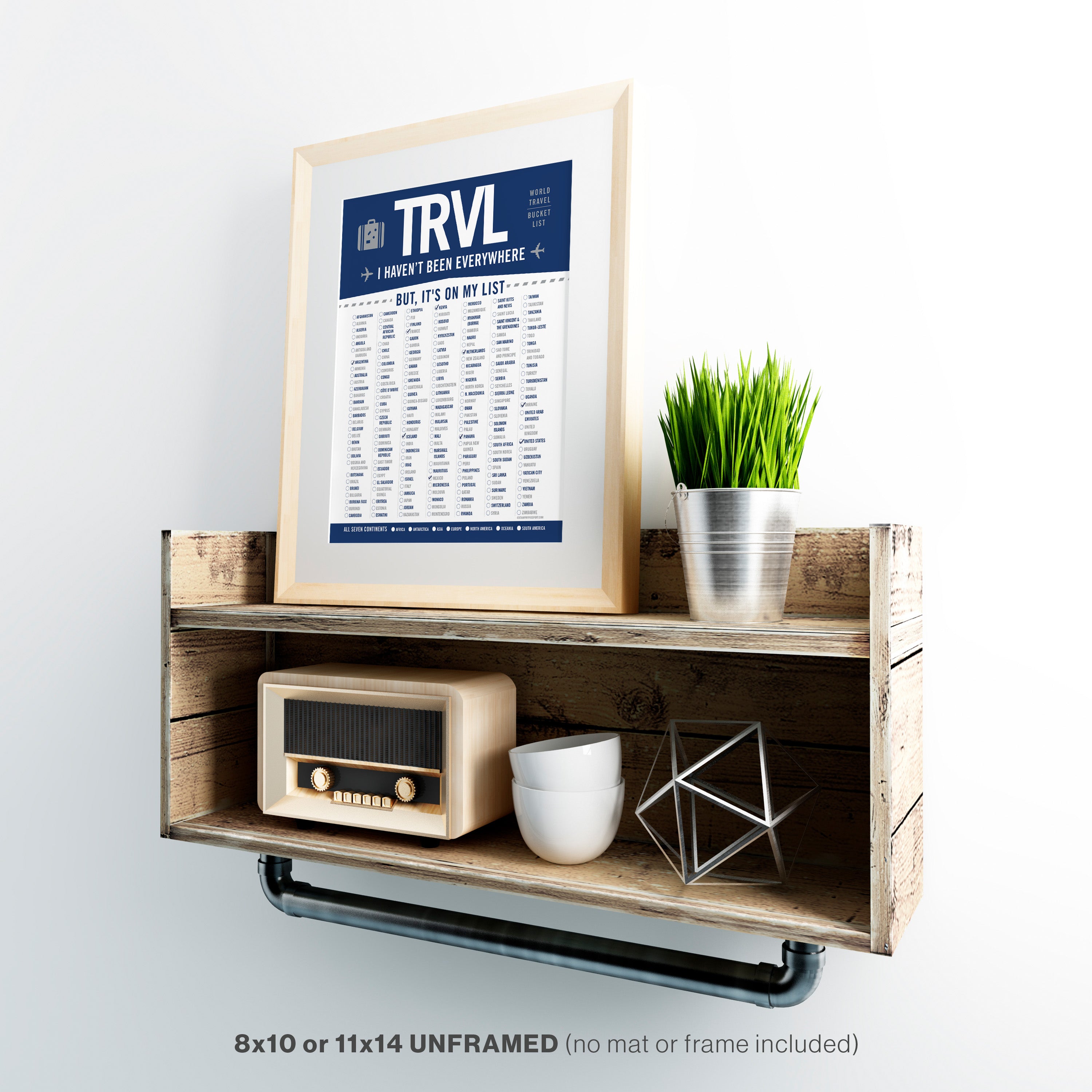 Framed world travel bucket list poster with 'TRVL' design on a wooden shelf with decorative items.